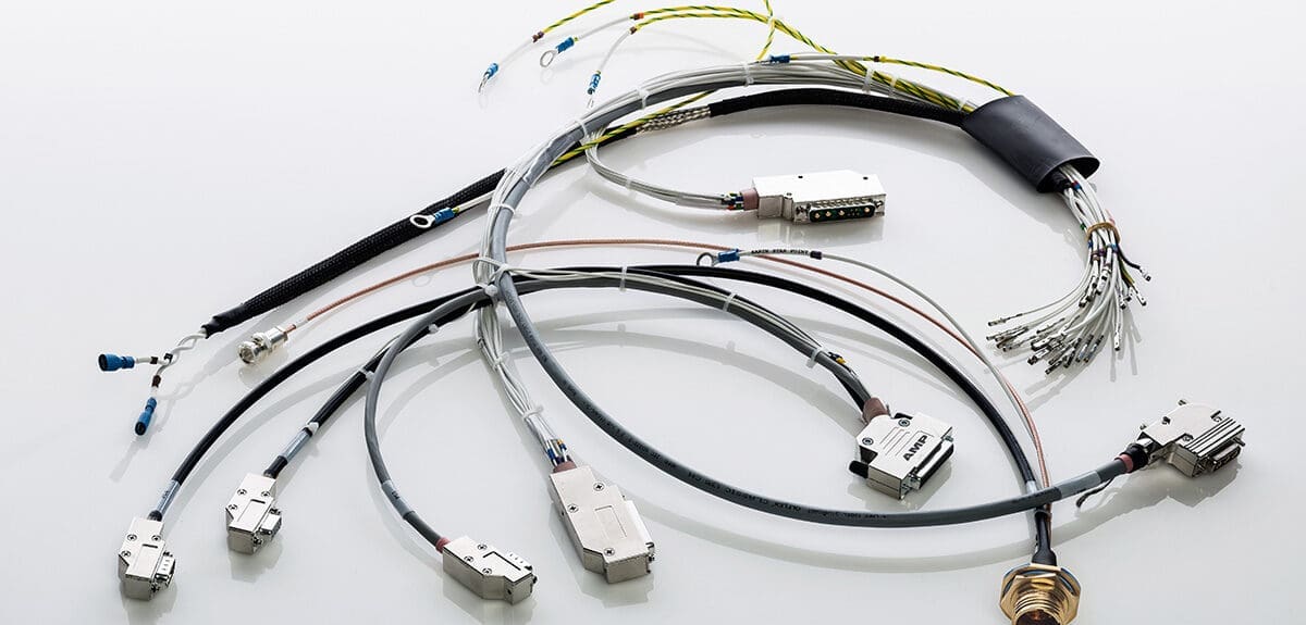 The image presents a variety of cables and electronic connectors arranged against a white background. In the foreground, there's a prominent circular connector, often seen in industrial or military applications, which can provide a secure and robust connection. There are several cables with D-sub connectors, commonly used for serial and video connections. Some cables have labels indicating specifications or destinations. A grouping of bare wire terminals is visible, likely intended for a screw or terminal block connection. The assortment of cables and connectors suggests that these are meant for a specialized electronic setup, such as a computer system, industrial machinery, or aerospace equipment, reflecting careful organization and the potential for a high level of complexity in the connected system.