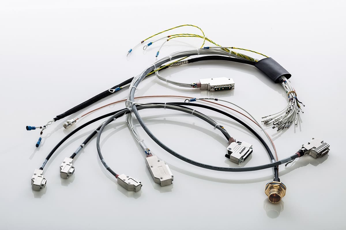 The image presents a variety of cables and electronic connectors arranged against a white background. In the foreground, there's a prominent circular connector, often seen in industrial or military applications, which can provide a secure and robust connection. There are several cables with D-sub connectors, commonly used for serial and video connections. Some cables have labels indicating specifications or destinations. A grouping of bare wire terminals is visible, likely intended for a screw or terminal block connection. The assortment of cables and connectors suggests that these are meant for a specialized electronic setup, such as a computer system, industrial machinery, or aerospace equipment, reflecting careful organization and the potential for a high level of complexity in the connected system.