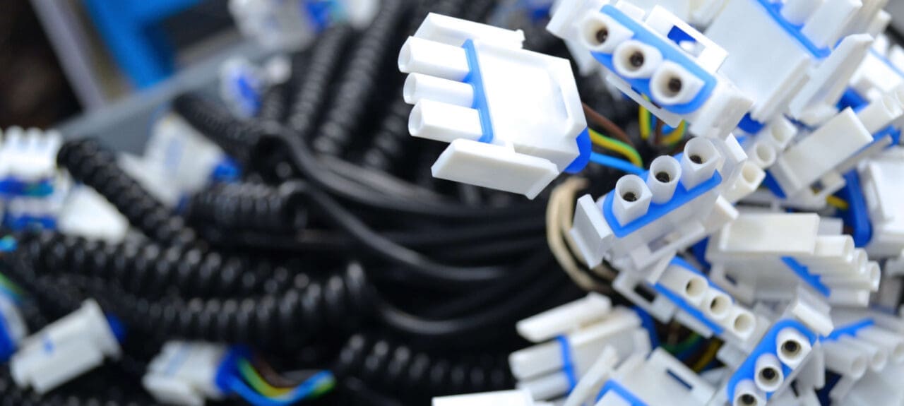 The image shows a large number of electrical connectors and coiled cables. The connectors are primarily white with blue accents, featuring multiple sockets, likely designed to accommodate spade or similar connectors. The black coiled cables suggest flexibility and potential use in environments where length variability is an advantage, like in telephone handsets or other electronic devices that require movement. The background is blurred with hints of blue, focusing the attention on the connectors in the foreground. The abundance and disarray of the connectors indicate a storage or supply setting, perhaps in an electronics workshop or manufacturing facility.