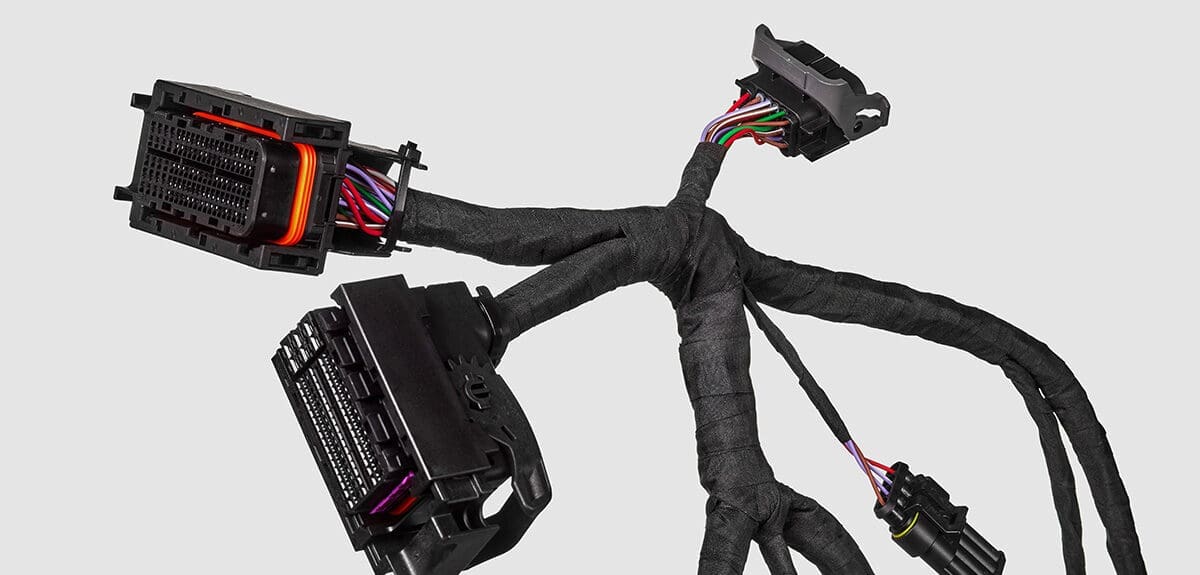 The image features an electrical wiring harness, which is commonly used in automotive and other complex machinery to bundle numerous wires together for organization and protection. The wires are encased in a protective black sheathing and split off into various branches, terminating in multi-pin connectors. The connectors are designed for quick release and secure connection, and the colored wiring within them is visible. Such harnesses are typically custom designed for specific applications, ensuring connectivity between different parts of a machine or vehicle. The grey background accentuates the harness, highlighting its intricate routing and construction.