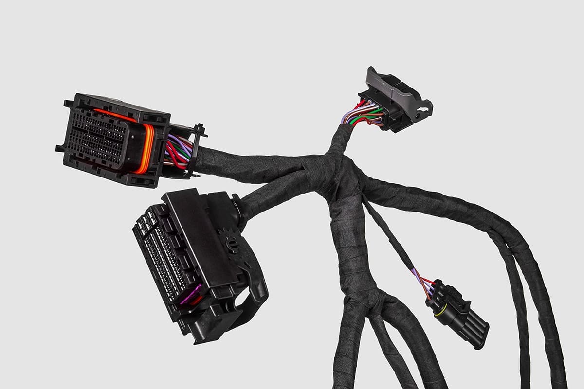 The image features an electrical wiring harness, which is commonly used in automotive and other complex machinery to bundle numerous wires together for organization and protection. The wires are encased in a protective black sheathing and split off into various branches, terminating in multi-pin connectors. The connectors are designed for quick release and secure connection, and the colored wiring within them is visible. Such harnesses are typically custom designed for specific applications, ensuring connectivity between different parts of a machine or vehicle. The grey background accentuates the harness, highlighting its intricate routing and construction.