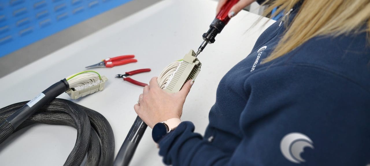 An employee at Cornelius Electronics is assembling a complex cable connector with precision tools on a workbench. Pliers and another cable lie beside her, and she's wearing a branded sweatshirt with the company logo.