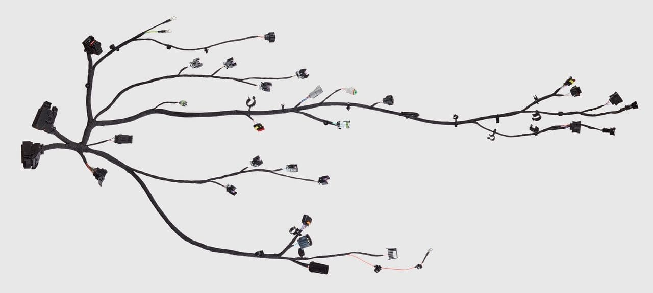 The image displays a complex array of black cables spread out against a light grey background. The cables branch out in different directions, resembling a tree or a river system with tributaries. Various connectors are attached at different points along the cables, with some ends exposed, indicating where they would connect to other devices. The connectors vary in size and shape, suggesting that they serve different functions or connect to different types of equipment. This setup is indicative of a wiring harness, which is typically used in automotive or electronic applications to organize and protect wires.