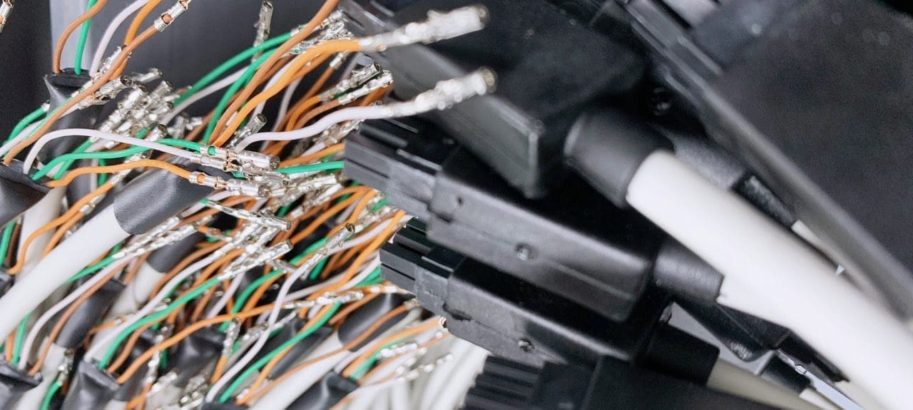 The image shows a close-up of a cluster of cables and connectors. We see a mix of shielded and unshielded wires, with various colours like orange, green, and white. Some of the wires are stripped at the end, exposing the metal conductors. These are typically used for making electrical connections in a system. A number of black connectors are visible, suggesting that these cables are part of a larger assembly or network of connections, possibly within a data center, telecommunications system, or complex electronic equipment. The focus is slightly blurred, adding a sense of depth and complexity to the array of connections.